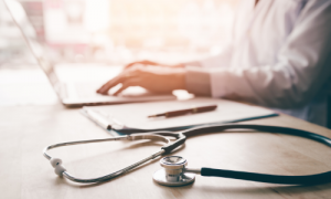 Doctor in background working on laptop with paper, pen, and stethescope laying on desk.