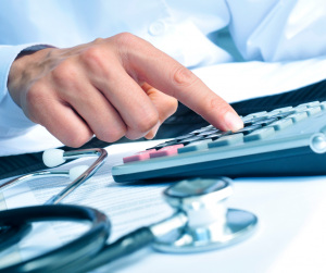 Stethascope sitting on table with doctors hand doing calculations on calculator.